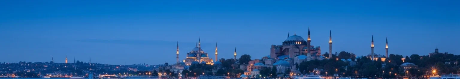 İstanbul'un simgesel Ayasofya ve Sultan Ahmet Camii'nin alacakaranlıkta aydınlatılmış, ön planda şehir ışıklarını yansıtan Boğaz'ın derin mavi gökyüzü altındaki çok geniş panoramik görünümü.